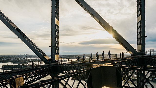 Six people walk along the gridwork at the top of the Sydney Harbour Bridge at sunrise.