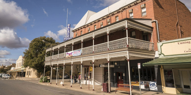  A three story red brick hotel with large early Victorian style white verandah across the front, framed by trees and blue sky. 