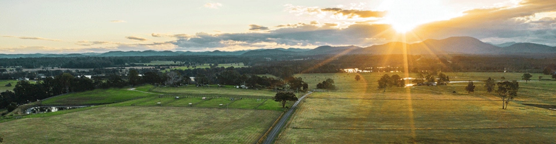 Top NSW road trips sunset over fields arial view of a road cutting through paddocks with silhouettes of hills under the setting sun in the distance