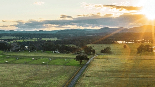 arial view of a road cutting through paddocks with silhouettes of hills under the setting sun in the distance