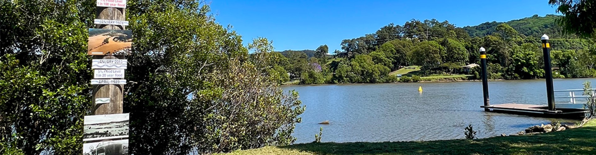 tweed-river-signpost-nsw-1920x500 A sunny, treed riverbank, with a pier and a wood post on shore with historical information, photos and swim warnings.