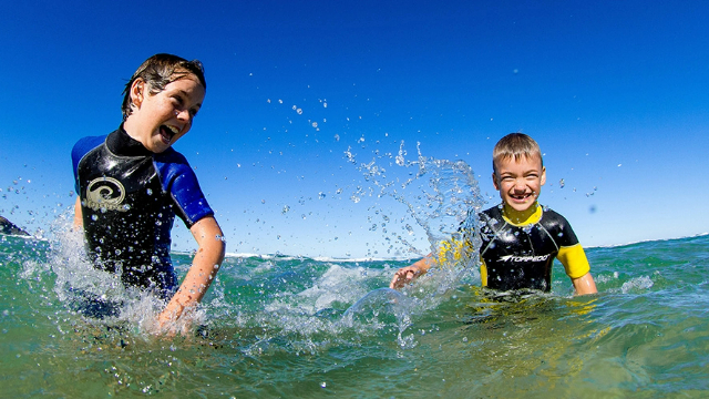 Two boys in rash guard shirts splash each other and laugh in waist deep ocean water, under blue sky.