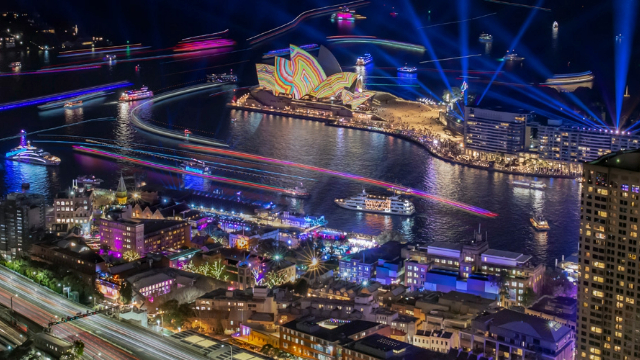 vivid-light-show-sydney-harbour-640x360 Sydney Harbour at night with the city and boats lit blue and purple, and a rainbow pattern over the Opera House.