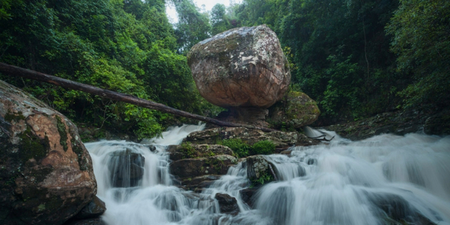  A small waterfall of rapids flows around a naturally stacked rounded rocks in the centre, surrounded by forest. 