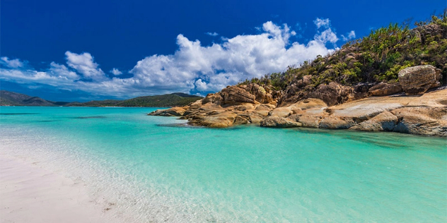  A bay of turquoise blue water framed by a rocky outcrop, white sand beach and green hills on the far shore. 