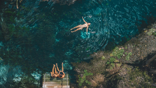 Aerial view of a woman in a floral bikini lit by the sun as she floats on her back by a grassy lakeshore. 