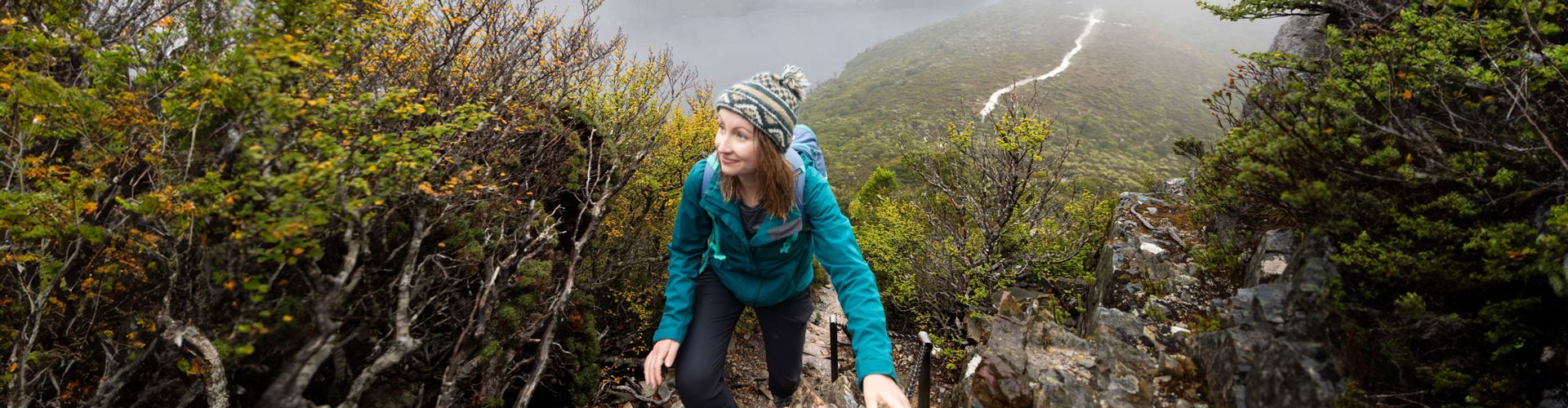 A woman in a blue hiking jacket climbs a mountain in Tasmania