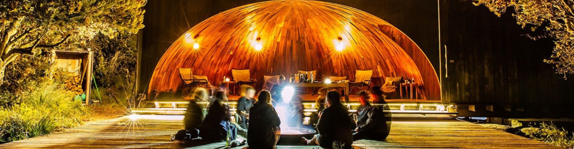 wukalina-walk-tasmania-1920x500 A group sits around a glowing firepit placed on a wooden boardwalk, flanked by trees and starry sky at Eddystone Point, Tasmania.