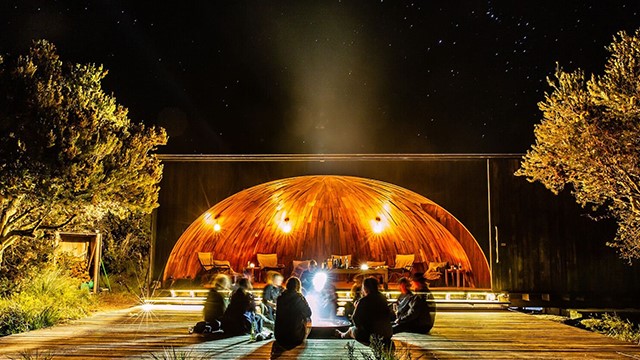 A group sits around a glowing firepit placed on a wooden boardwalk, flanked by trees and starry sky at Eddystone Point, Tasmania. 
